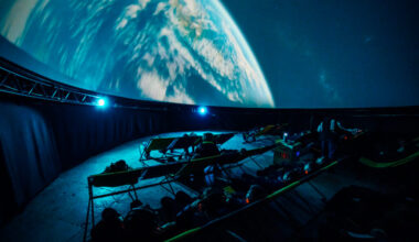 The inside of a planetarium in the dark with an image of the moon on the curved roof and deck chair seating below