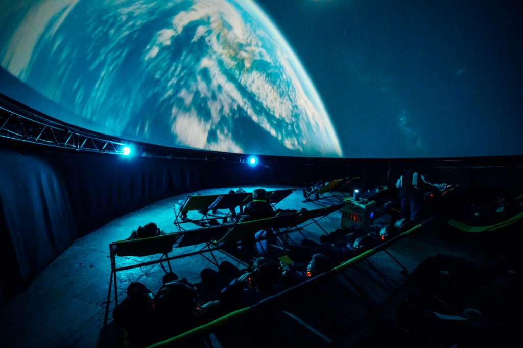 The inside of a planetarium in the dark with an image of the moon on the curved roof and deck chair seating below