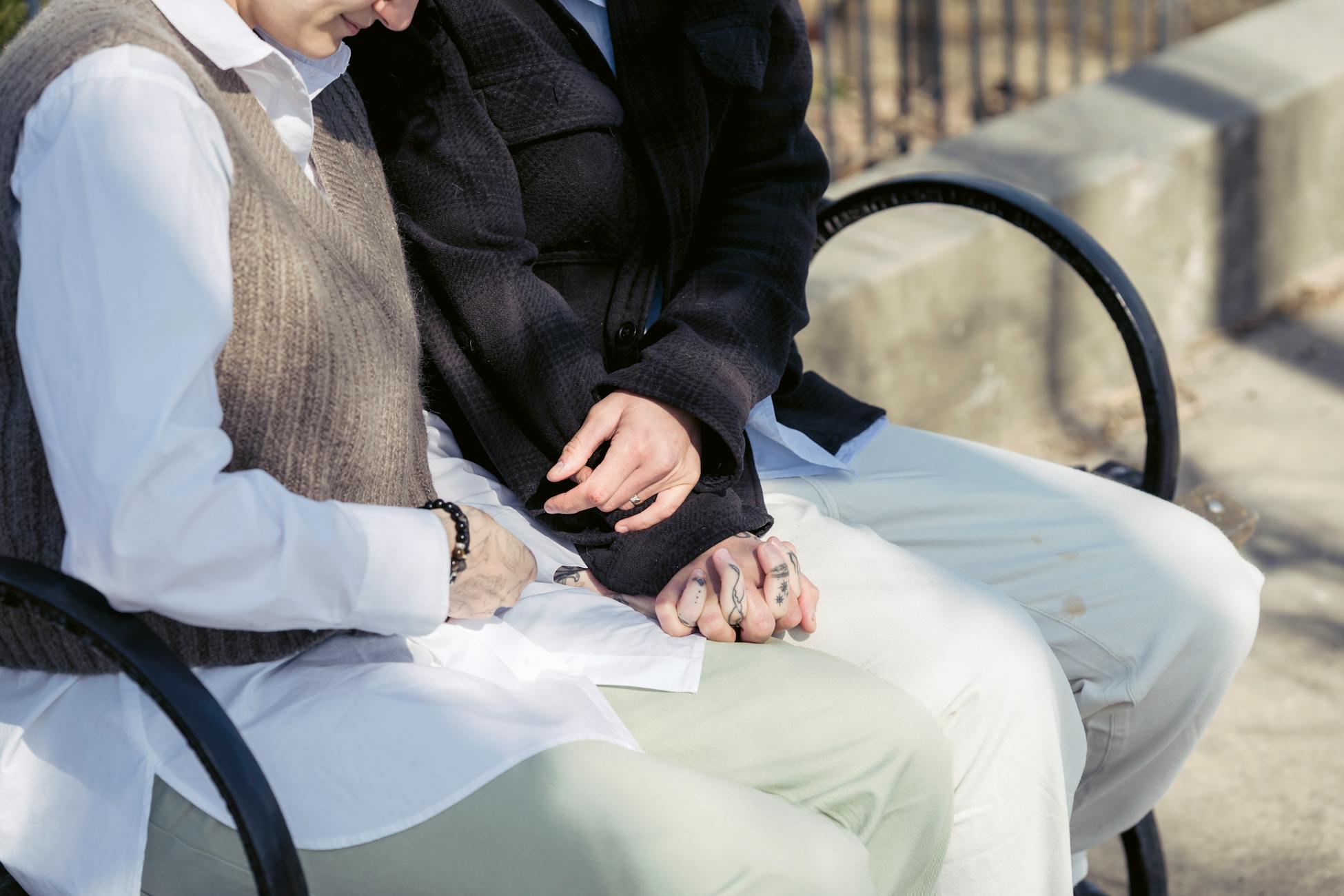 Crop unrecognizable homosexual woman with tattoo holding beloved by hand while sitting on bench in sunny town
