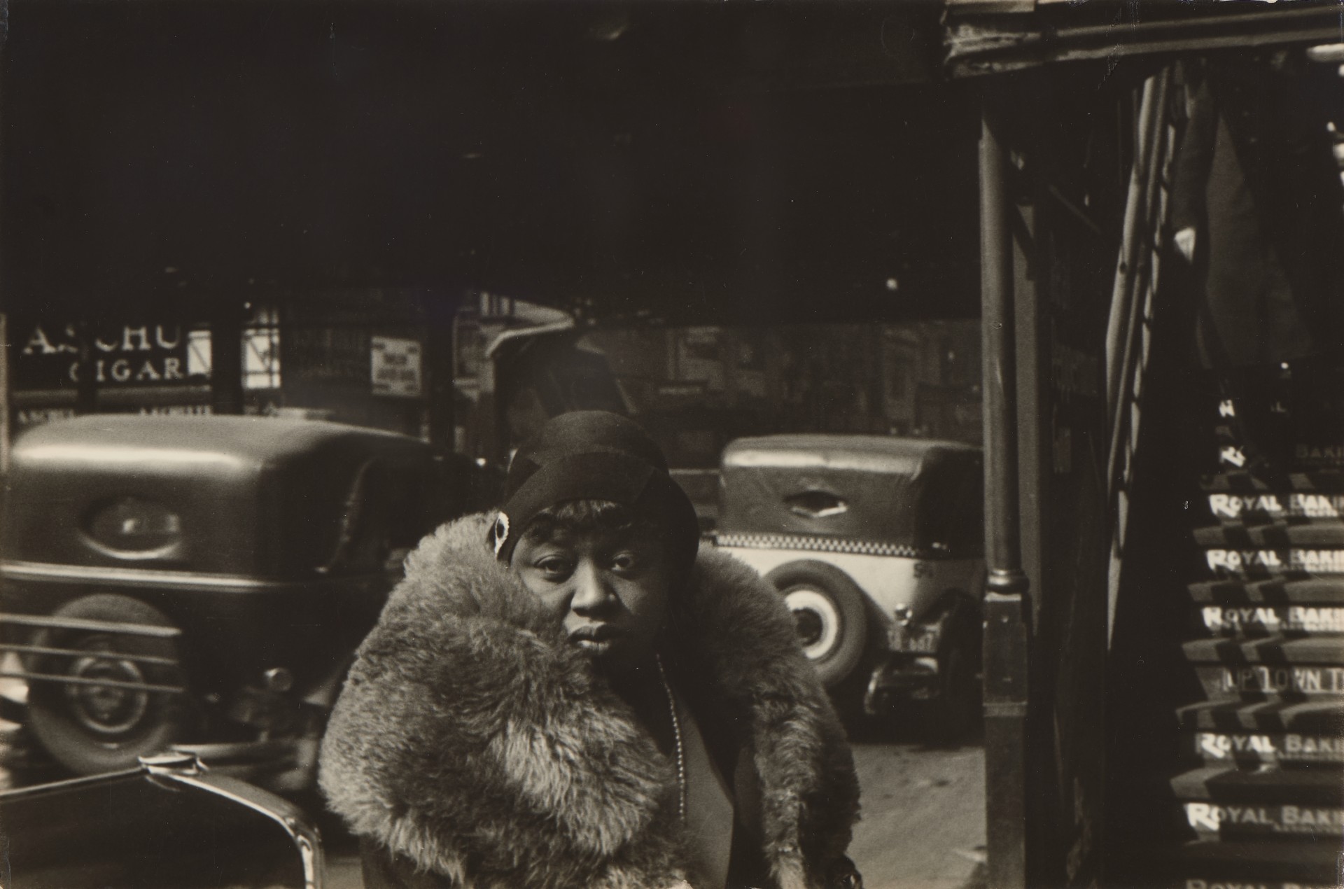 A woman in a large fur collar turns to look at the camera amid the noise and clutter of a night-time New York street, taxis and a cigar-store sign visible behind her.