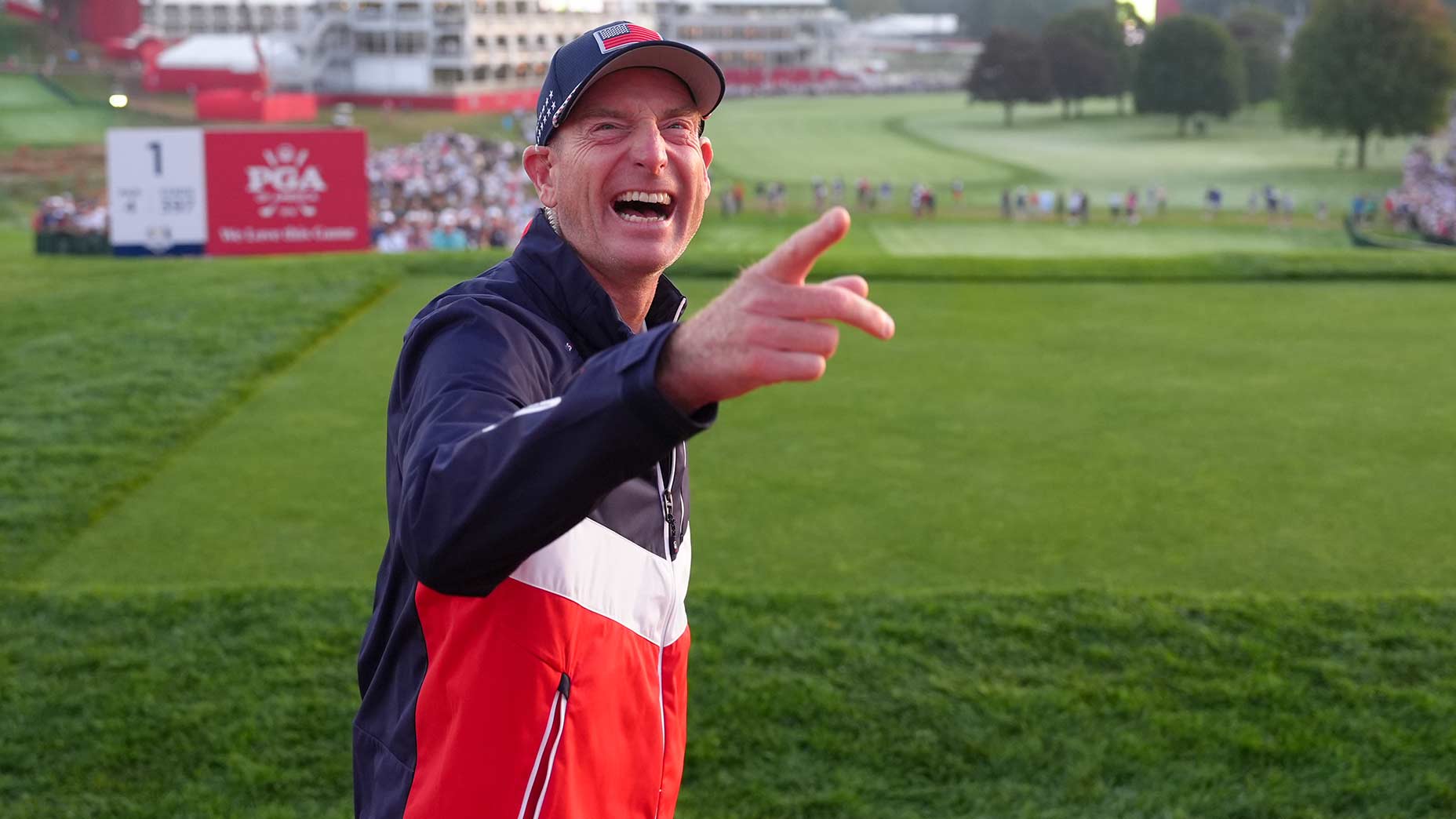 Ryder Cup captain Jim Furyk, wearing a red, white, and blue jacket and cap, smiles and points while standing on a golf course near a PGA sign, with spectators and trees in the background.