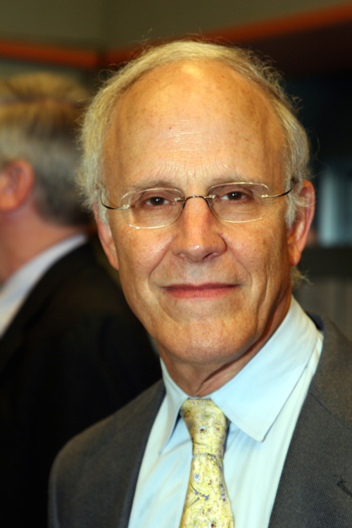 A man with white hair and glasses wearing a gray blazer, blue button up shirt and yellow tie looks at the camera.