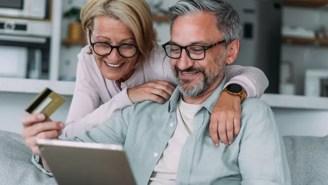 A happy older couple at home together, using tablet for online shopping.