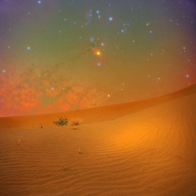 View over warmly lit dunes with glowing, billowing Milky Way near the horizon and many colorful stars above.