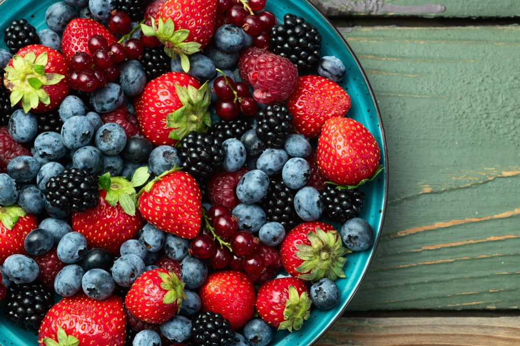 A mix of ripe, colorful berries in a turquoise bowl, including blueberries, strawberries, raspberries, blackberries, and red currants, on a rustic wooden background.