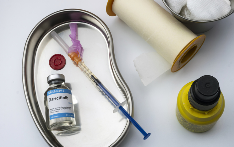 A close up of a clear glass bottle labeled baricinitib, with a syringe next to it. Next to the bottle and syringe is medical tape and other bottles.