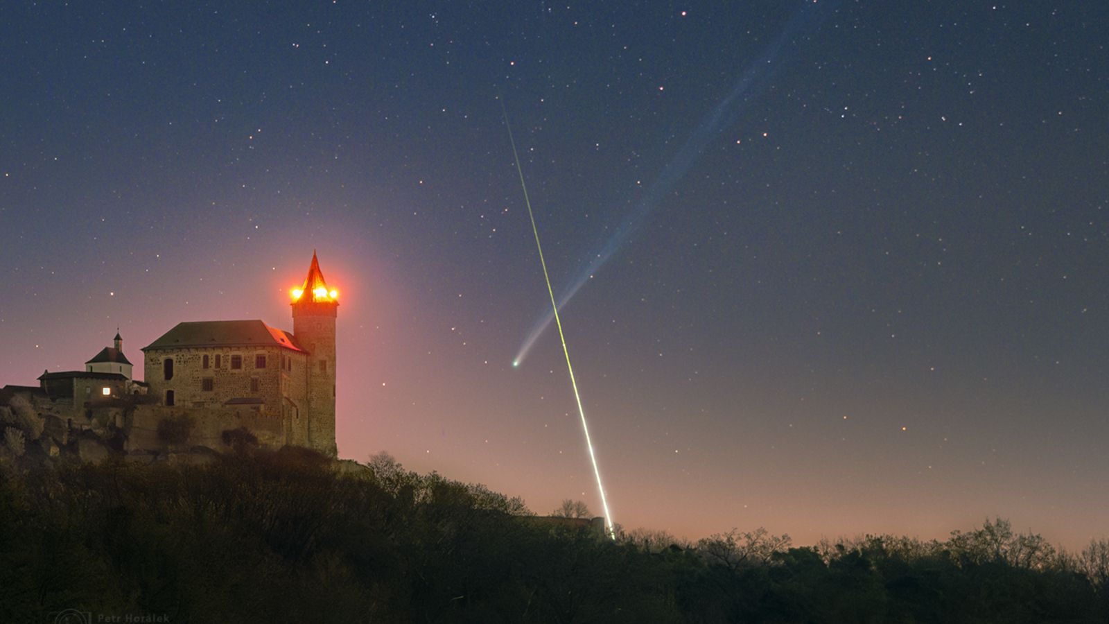 A timelapse image showing a bright streak of light crossing in front of a comet's long tail to form an X-like shape in the night sky. A medieval castle is illuminated in the foreground.