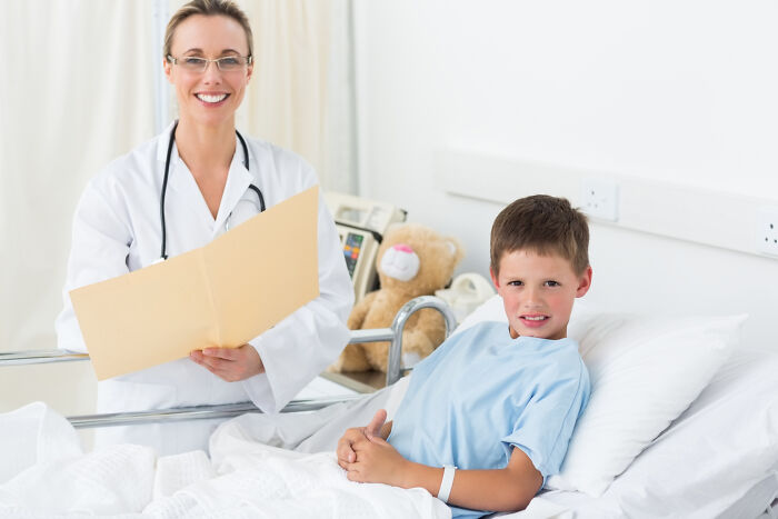 Smiling surgical staff reviewing patient files with a young boy in hospital bed during sedation recovery.