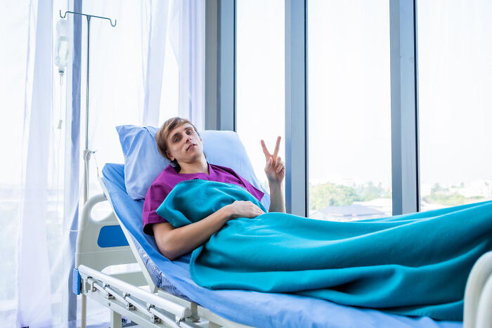 Young surgical staff resting in hospital bed covered with blue blanket near large window after sedation.