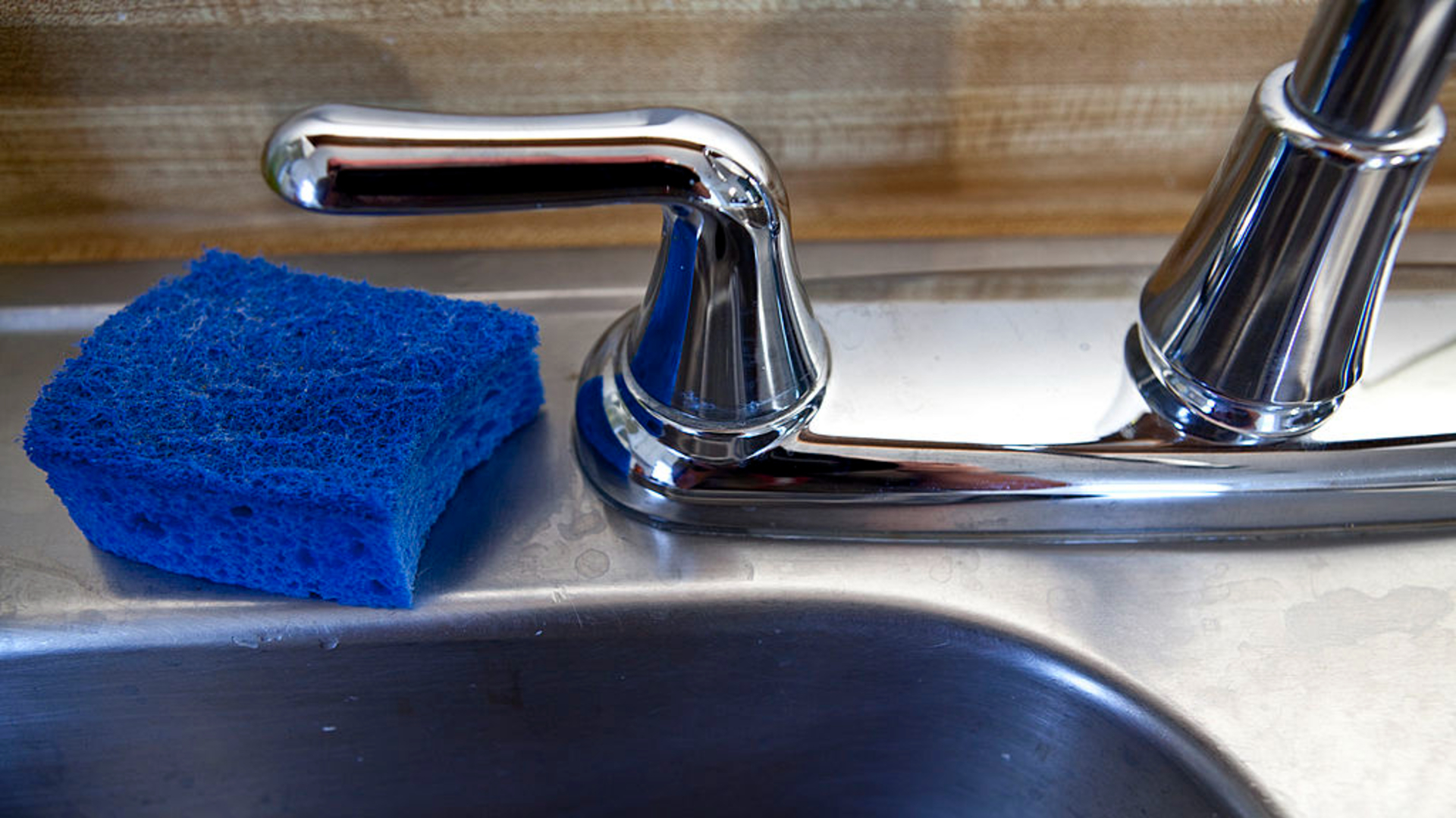 A blue square dish sponge sits to the left of a metal sink
