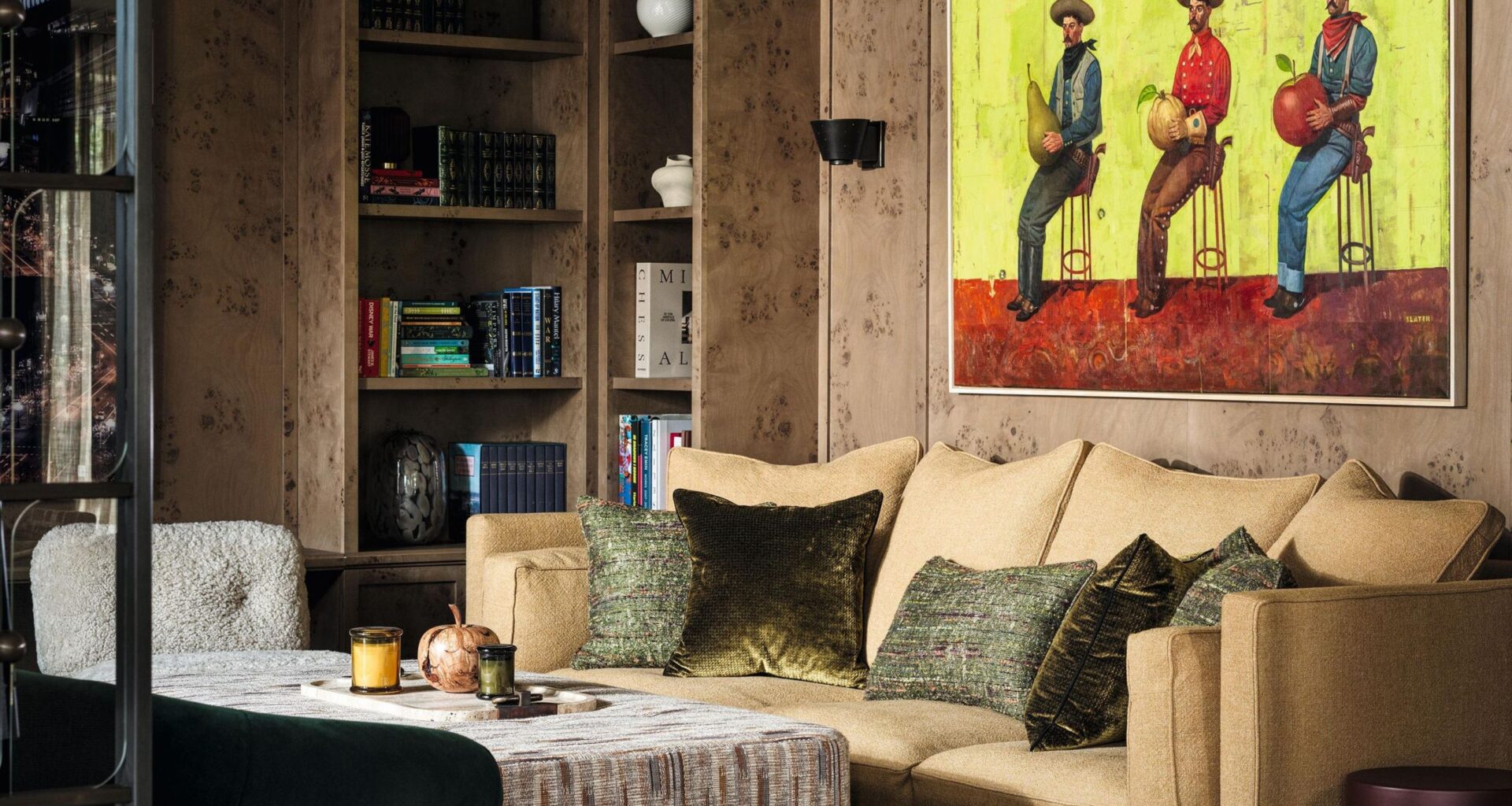 a burl wood clad sitting area with built in shelving, a beige couch and a painting of cowboys on the wall