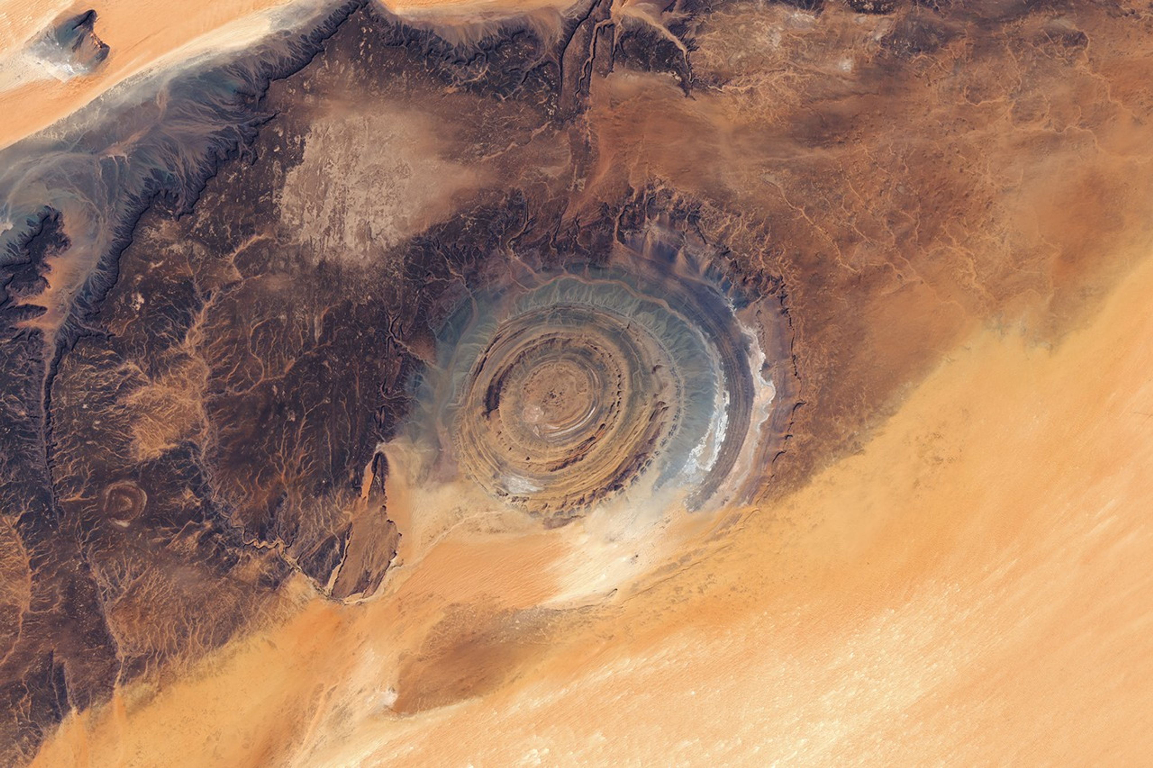 A large "bull's-eye" feature composed of concentric rock formations of various colors is imprinted on the edge of a dark plateau flanked by desert sands.