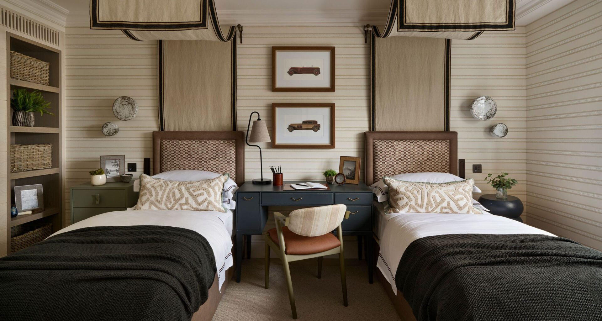 a twin children's bedroom with beige striped walls and two canopy twin beds, a desk in the middle, a bookcase, and dark green blue color pops