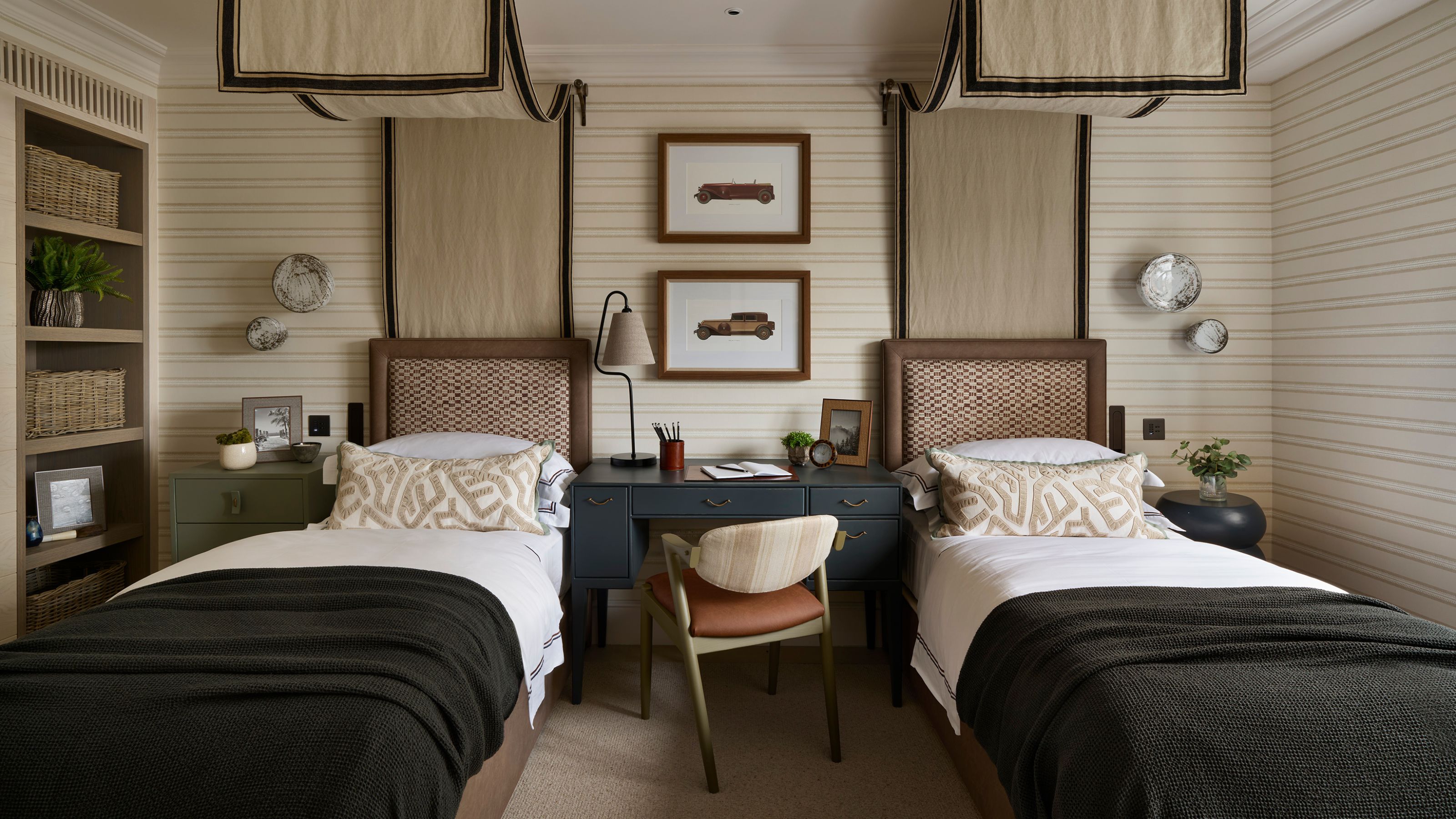 a twin children's bedroom with beige striped walls and two canopy twin beds, a desk in the middle, a bookcase, and dark green blue color pops