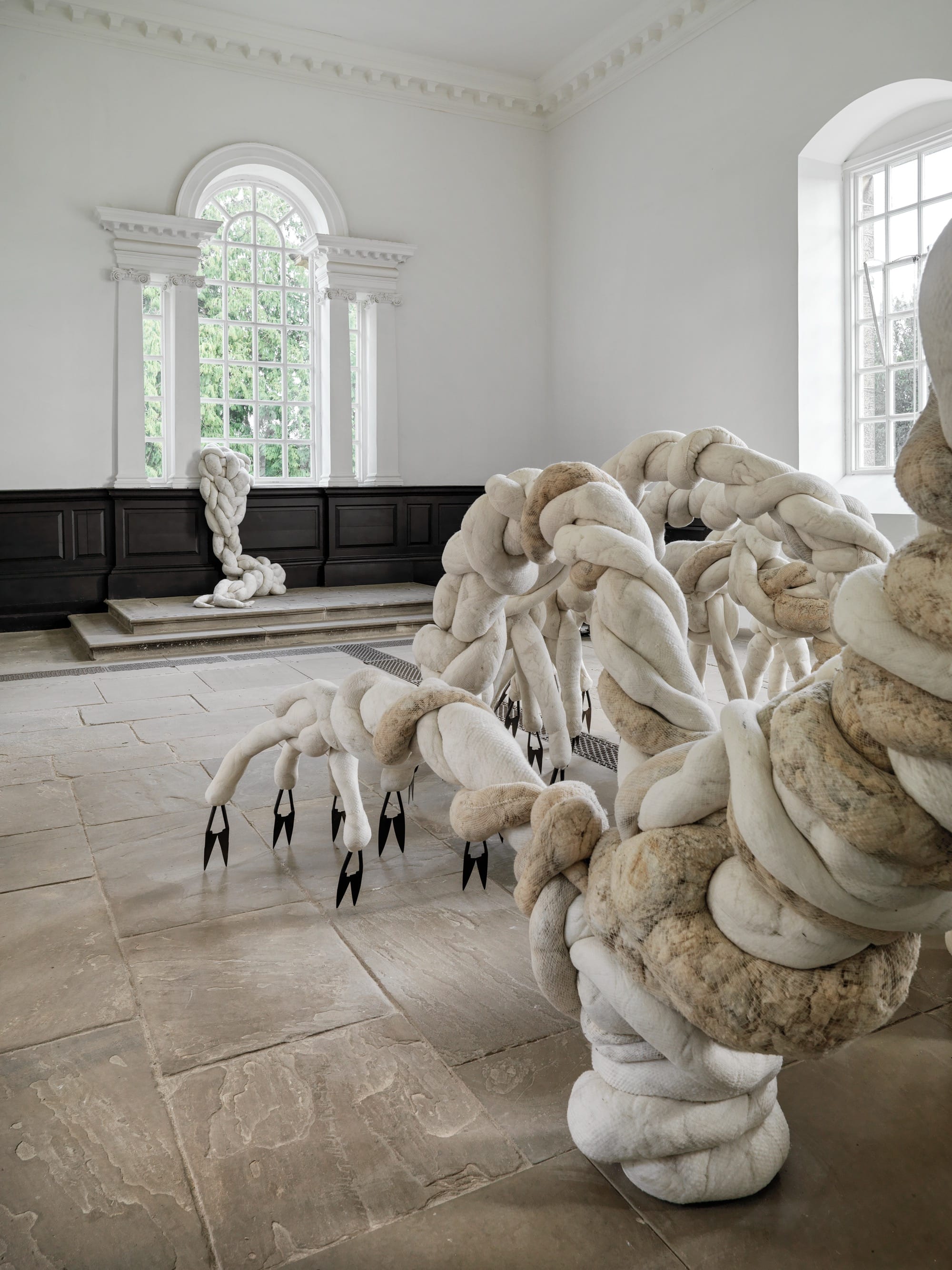 a large-scale installation by Nicola Turner inside of Yorkshire Sculpture Park's 18th-century chapel gallery space of a textile form that appears to be crawling or expanding across the room, out of an opening toward the ceiling, with sharp clippers and scythes for "feet"