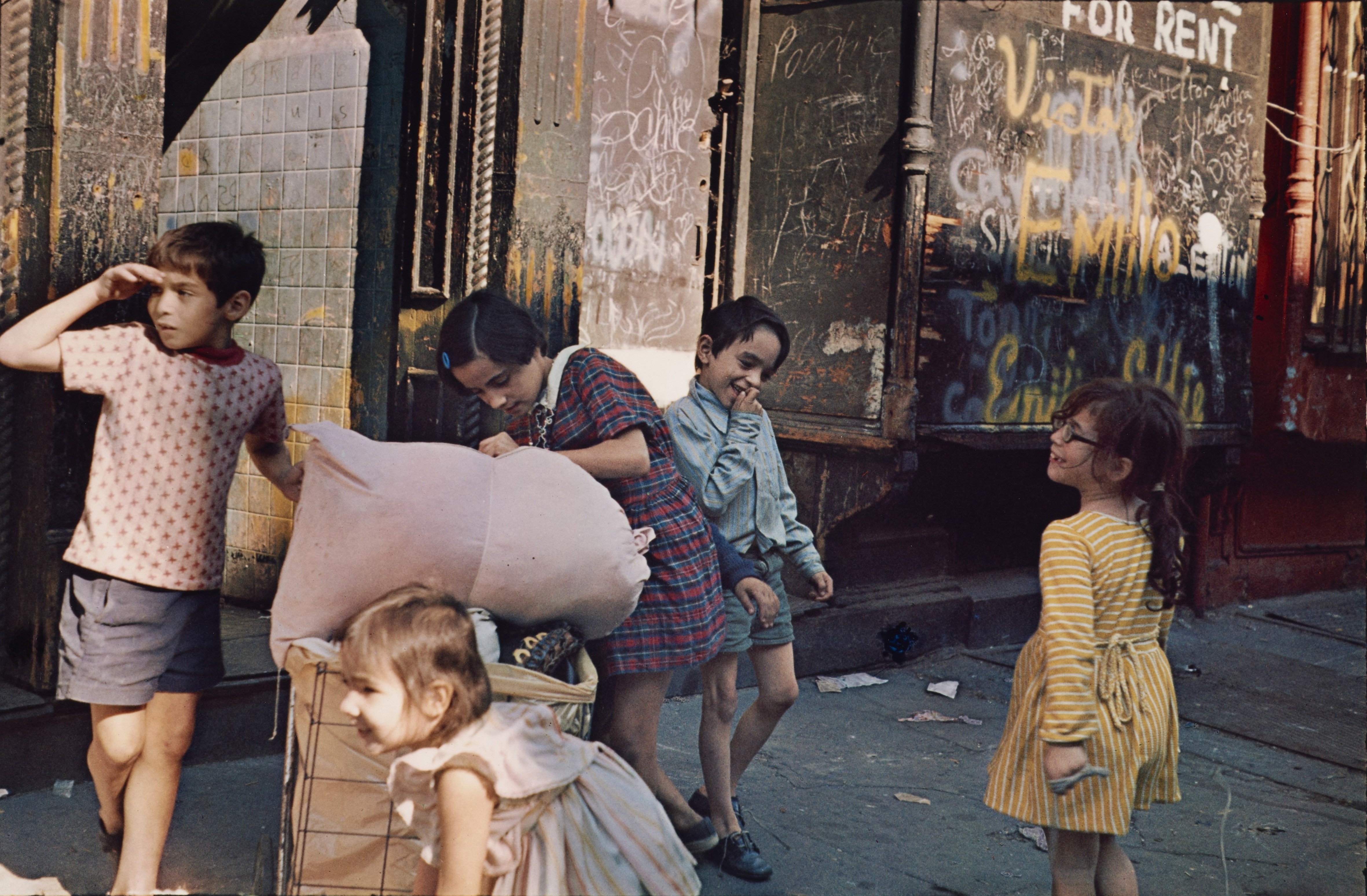 Five children laugh and jostle on a sunlit pavement outside a row of derelict, graffiti-covered shopfronts, one toddler sitting in a wire cart loaded with a pink bundle.