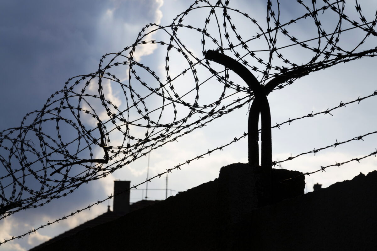 Photo shows a roll of barbed wire at the top of a prison wall. UK prisons are now required to take imprisoned vegan people’s ethical beliefs into account while planning menus.