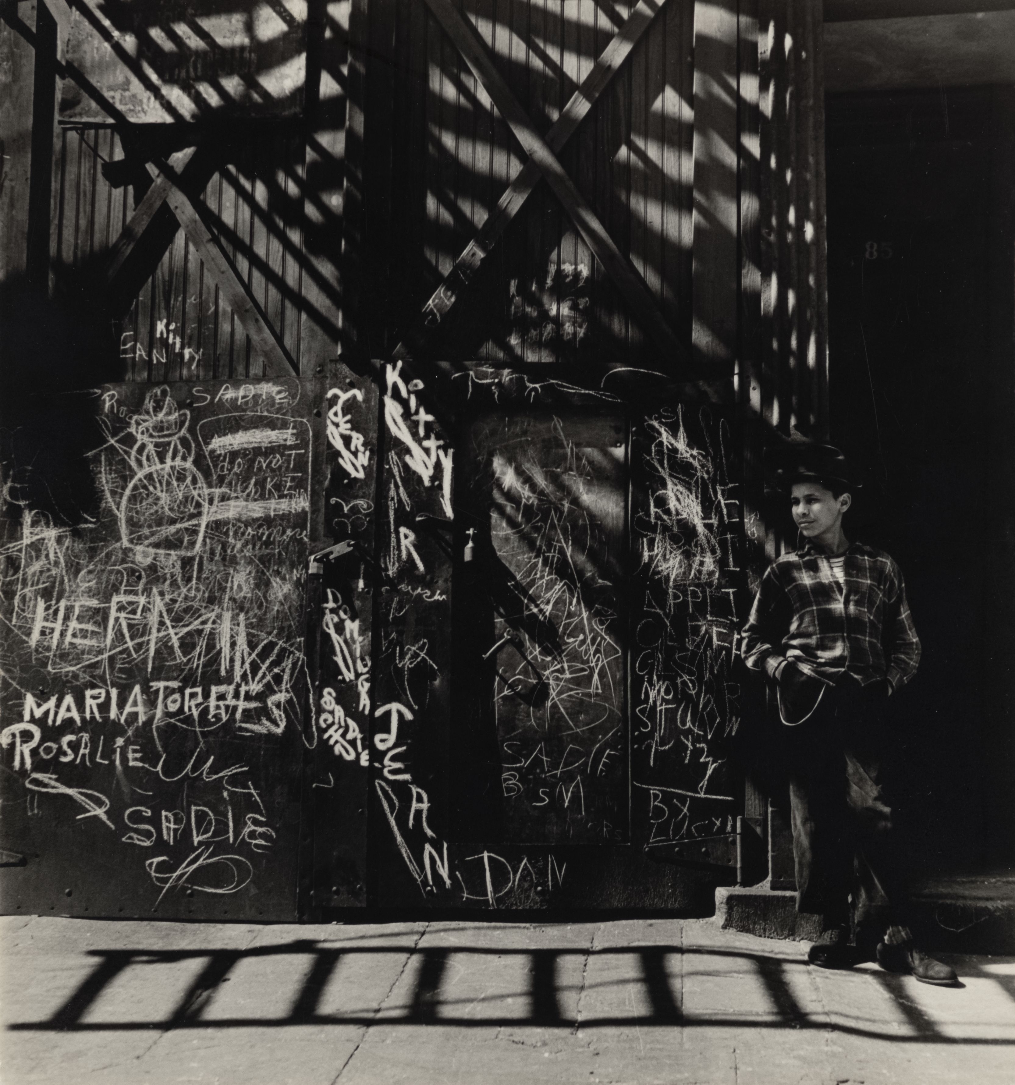 A teenage boy in a checked jacket leans against a wooden door covered in chalked names and graffiti, his arms folded, half in deep shadow cast by a fire escape overhead.