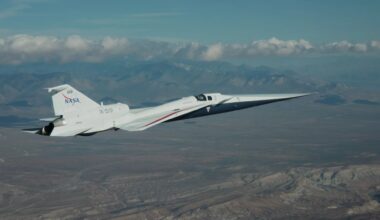 NASA’s X-59 flies above the Mojave Desert on a clear day. The white aircraft has light gray, red, and blue accents, with a NASA logo and the number 859 on its tail. It appears flying level over the desert landscape, with a mountain range visible on the horizon and a trail of clouds above.