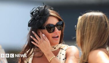 A woman with dark hair wears a white and black formal polka-dot dress, with a black fascinator and large black sunglasses - she is talking to a woman with blonde hair, and both are outside