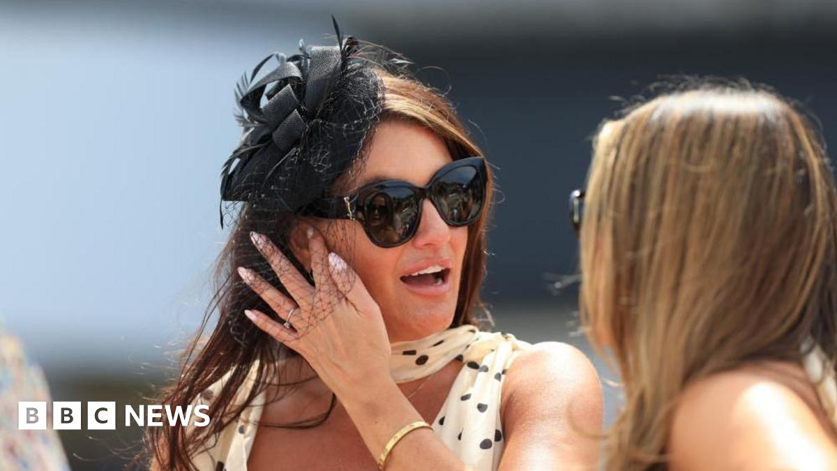 A woman with dark hair wears a white and black formal polka-dot dress, with a black fascinator and large black sunglasses - she is talking to a woman with blonde hair, and both are outside