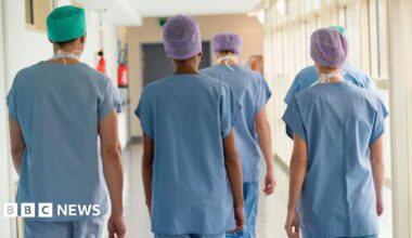 Five medical staff members walk through a hospital corridor. Their backs are to the camera. They are wearing blue medical scrubs and paper hats.