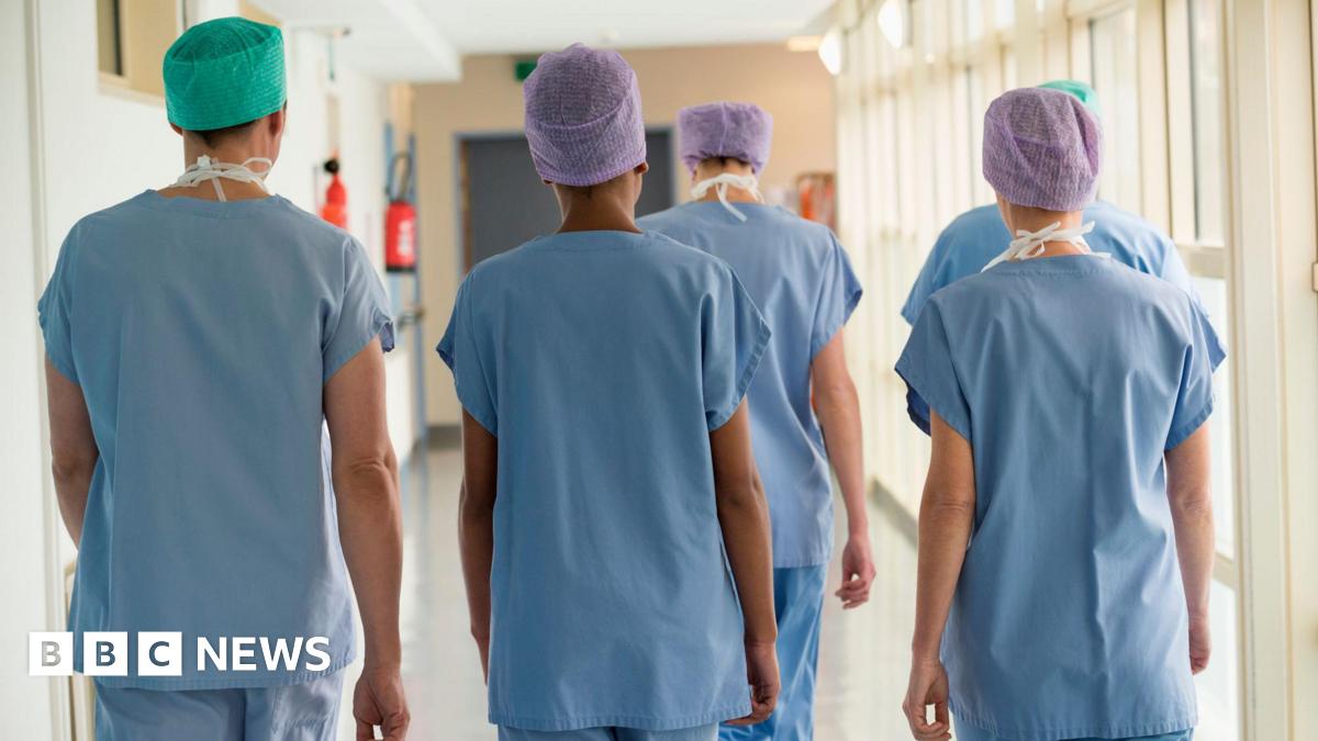 Five medical staff members walk through a hospital corridor. Their backs are to the camera. They are wearing blue medical scrubs and paper hats.