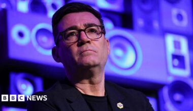 Andy Burnham looks serious while standing in front of a blue and purple background.