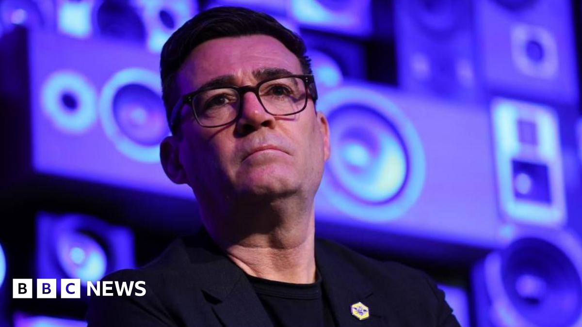 Andy Burnham looks serious while standing in front of a blue and purple background.