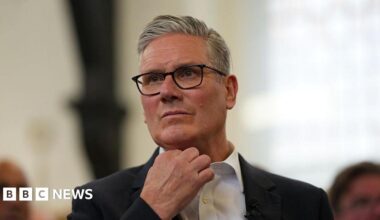 Sir Keir Starmer, with short grey hair and black glasses, stands up while wearing a white shirt with a black suit jacket. The background behind him is blurred.