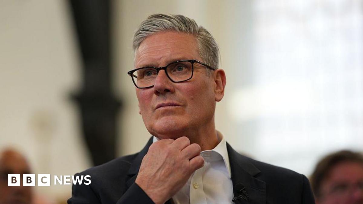 Sir Keir Starmer, with short grey hair and black glasses, stands up while wearing a white shirt with a black suit jacket. The background behind him is blurred.