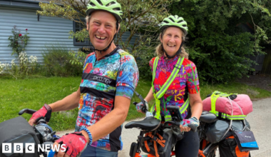 Chris and Nicky - dressed in multi-coloured cycling T-shirts and neon green helmets - are sitting on their orange tandem, which has luggage on the back. They are on a country road with greenery and a pale blue-painted house, partially visible, in the background