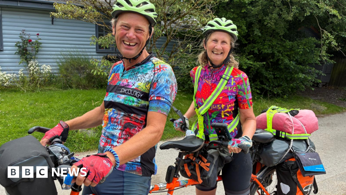 Chris and Nicky - dressed in multi-coloured cycling T-shirts and neon green helmets - are sitting on their orange tandem, which has luggage on the back. They are on a country road with greenery and a pale blue-painted house, partially visible, in the background