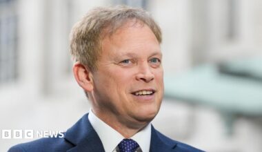 Grant Shapps has fair hair and is wearing a navy jacket over a white shirt and blue and pink tie. He is smiling with his mouth open.