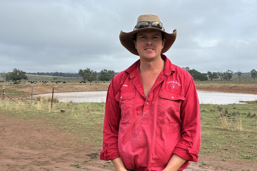 A man in a red shirt and a hat stands in front of a dam.  