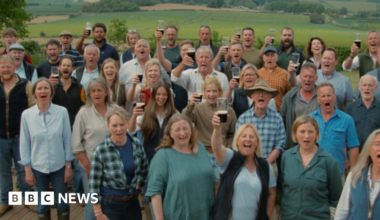 The choir is standing outside and looking at the camera, many toasting the picture with a pint. There are men and women with Jeremy Clarkson in the middle.