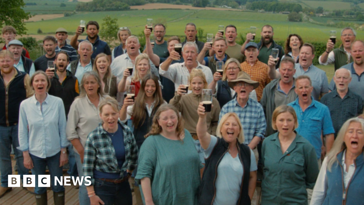 The choir is standing outside and looking at the camera, many toasting the picture with a pint. There are men and women with Jeremy Clarkson in the middle.