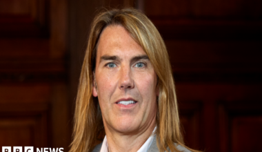 A man with long golden hair looking into the camera wearing a grey suit and white shirt.