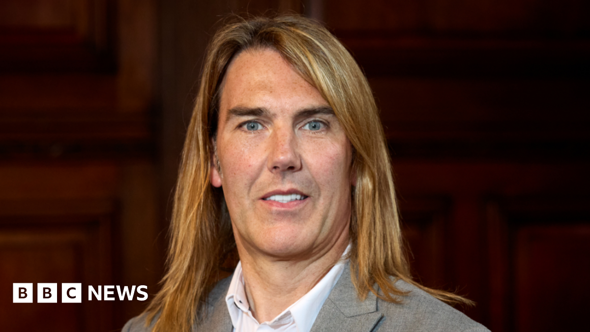 A man with long golden hair looking into the camera wearing a grey suit and white shirt.