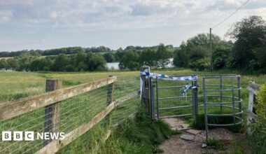 Blue and white police tape on a kissing gate style fence. In the background are fields, trees and a large body of water visible between trees.