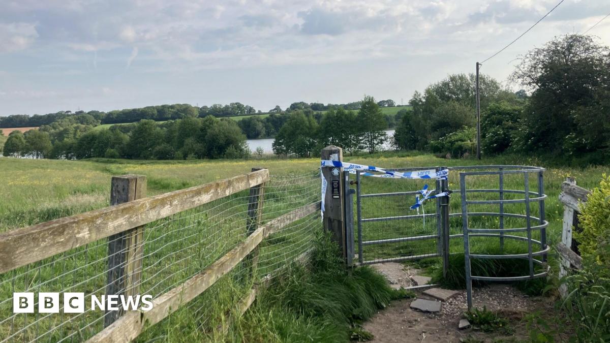 Blue and white police tape on a kissing gate style fence. In the background are fields, trees and a large body of water visible between trees.
