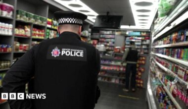 A police officer crouching down behind the counter of a mini-mart where there is a hidden compartment in the shelving, which he has opened and is looking into.