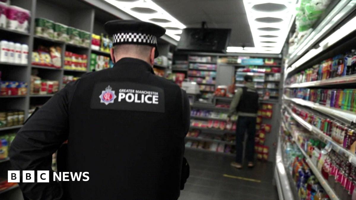A police officer crouching down behind the counter of a mini-mart where there is a hidden compartment in the shelving, which he has opened and is looking into.