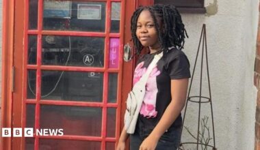 A girl is smiling while standing next to a red phone box. She has a black and pink Barbie T-shirt, jeans and a white bag.