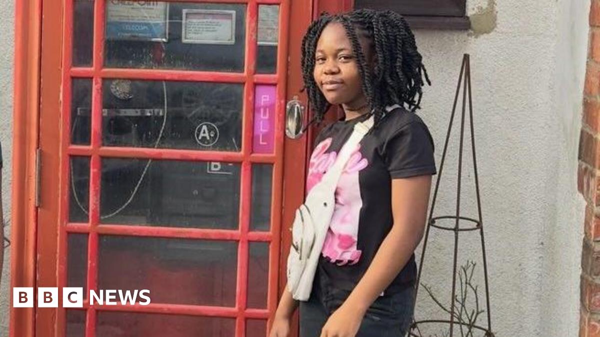 A girl is smiling while standing next to a red phone box. She has a black and pink Barbie T-shirt, jeans and a white bag.