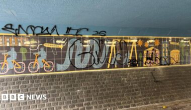 Art on the wall of an underpass, which depicts colourful cartoon figures riding bikes and playing on swings against a black background. However, a large graffiti tag has been sprayed over the top.