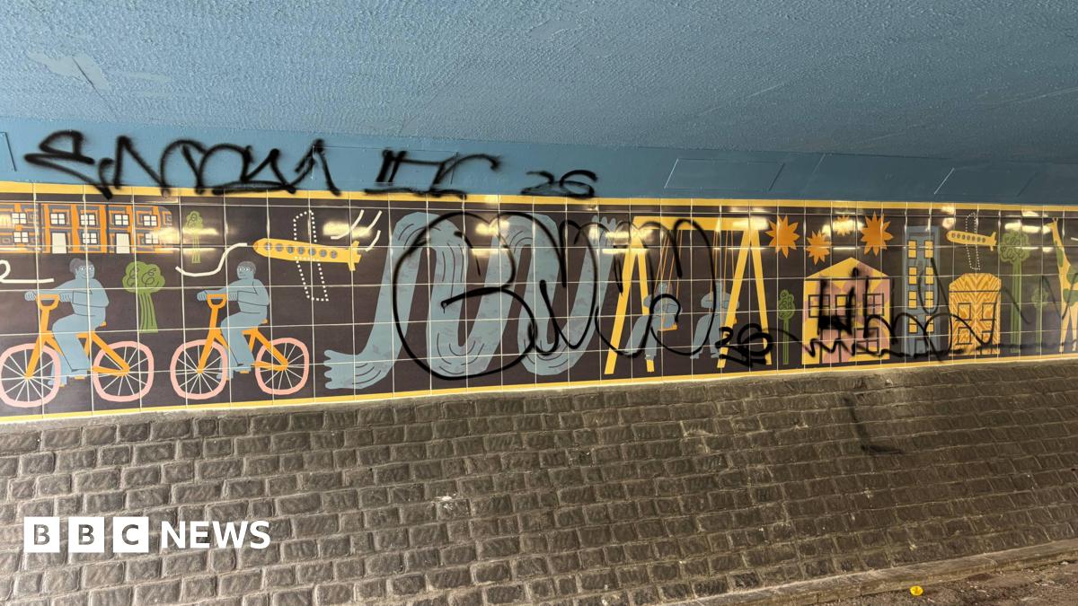 Art on the wall of an underpass, which depicts colourful cartoon figures riding bikes and playing on swings against a black background. However, a large graffiti tag has been sprayed over the top.