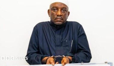 Saleh Mamman wearing a black cloth, handcuffed and holding a white board