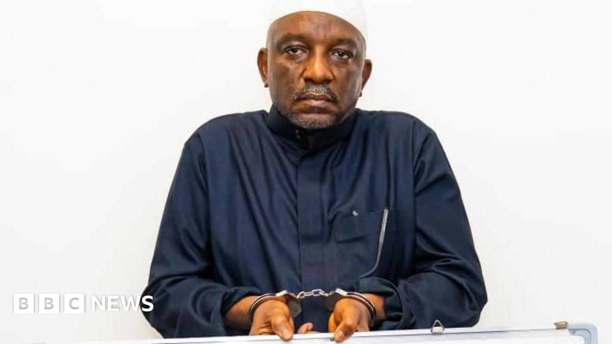 Saleh Mamman wearing a black cloth, handcuffed and holding a white board