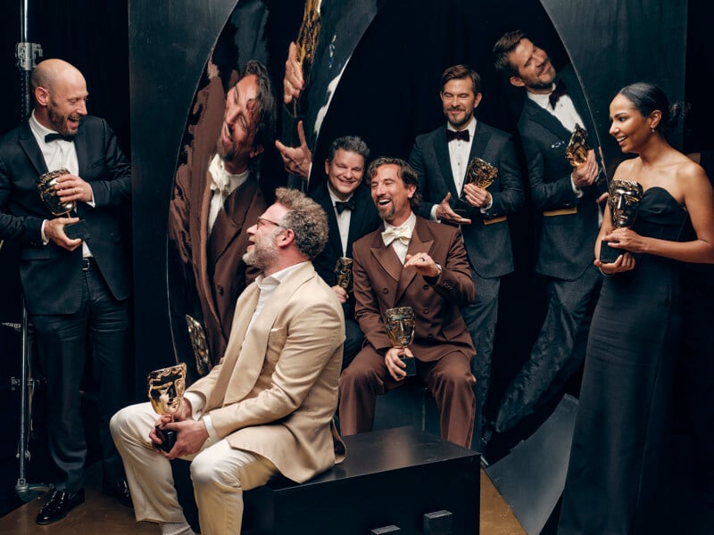 Six well-dressed people holding awards, laughing and posing together in front of mirrored panels; five stand while one in a beige suit sits, reflecting a celebratory and joyful atmosphere.
